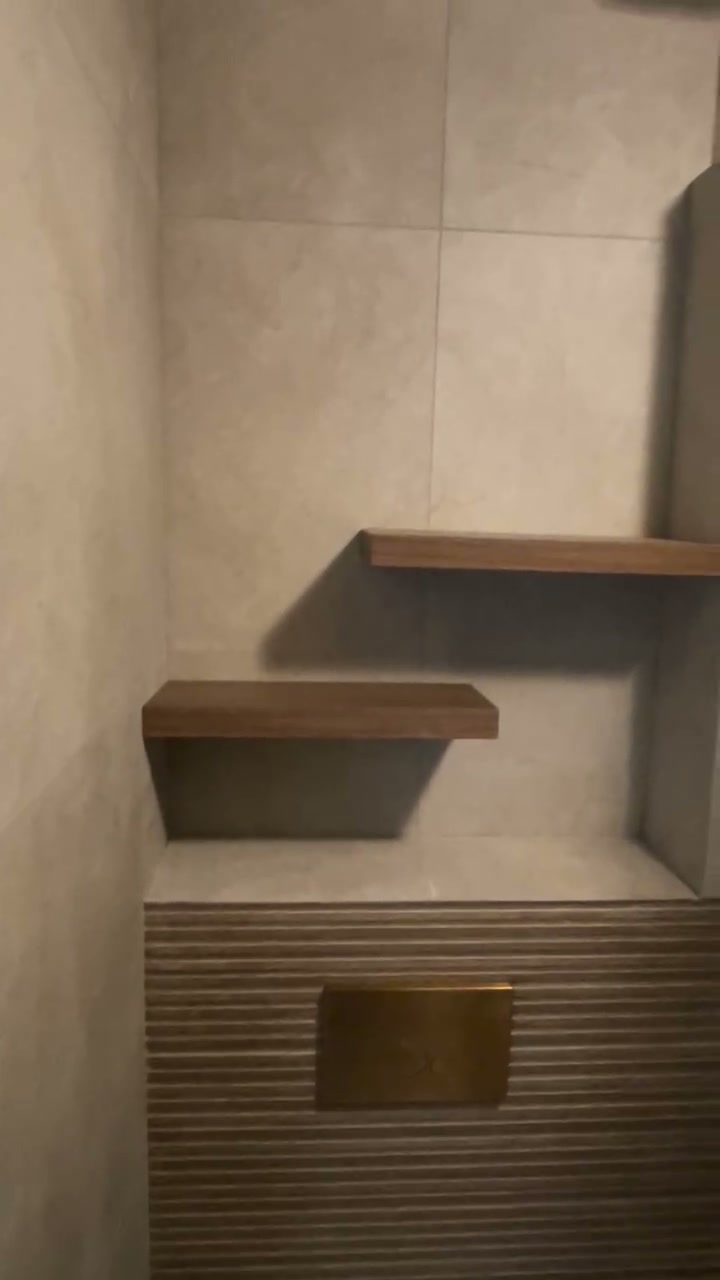 Toilet met een geribbelde tegelwand, een koperen bedieningsplaat en twee houten wandplanken