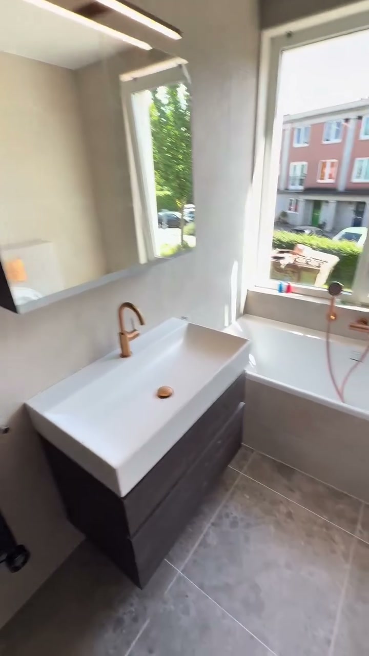 Wit wastafelmeubel met een koperen kraan en spiegelkast, naast een ligbad onder het raam